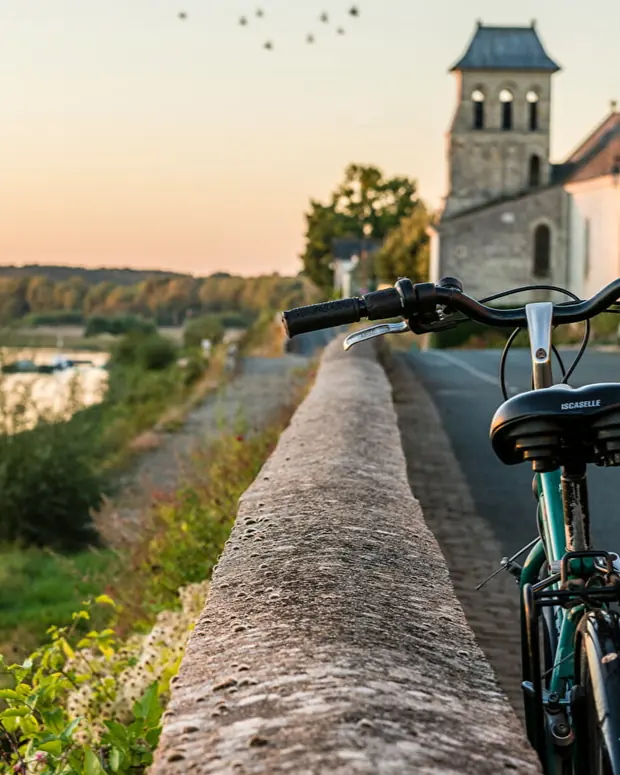 Un vélo posé contre le muret d'un chemin longeant la Loire, une église de village au loin au coucher du soleil