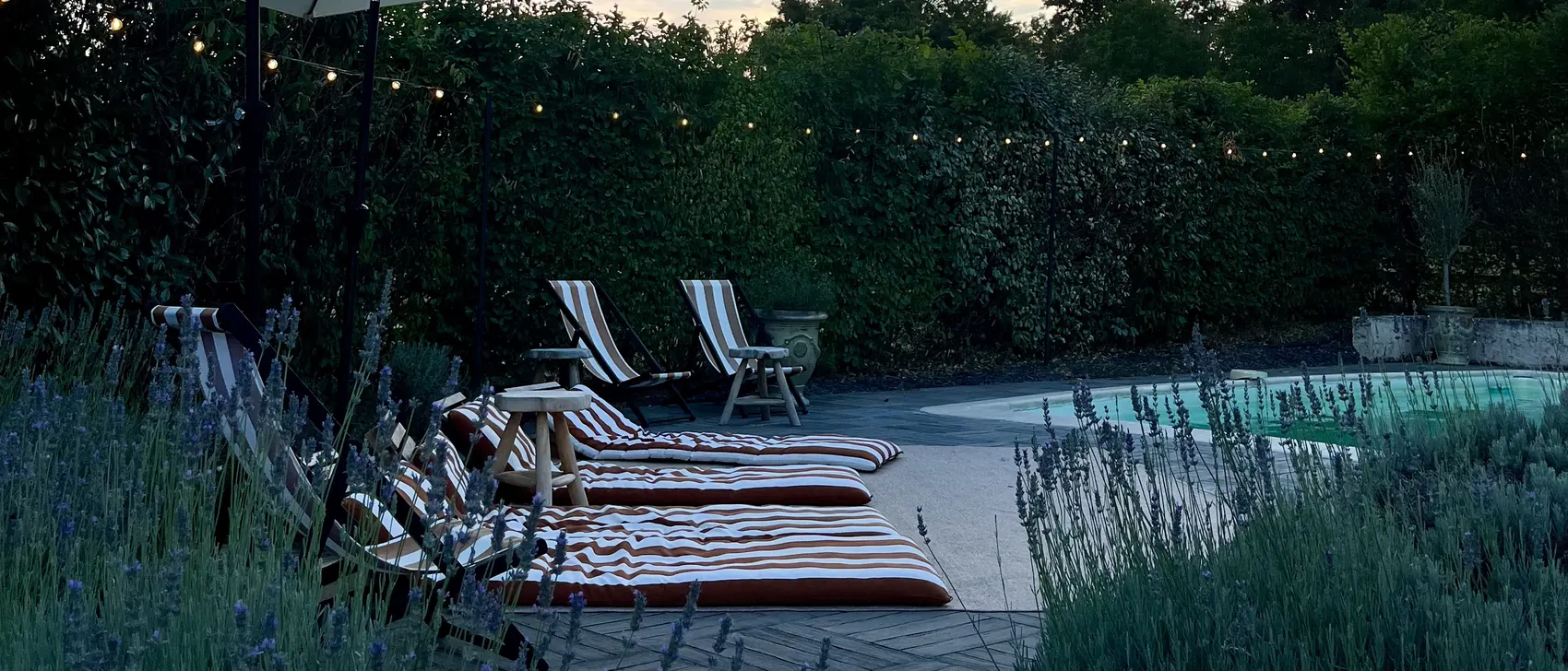 La piscine au crépuscule, ses transats rayés alignés le long des haies de lavande, sous les guirlandes lumineuses